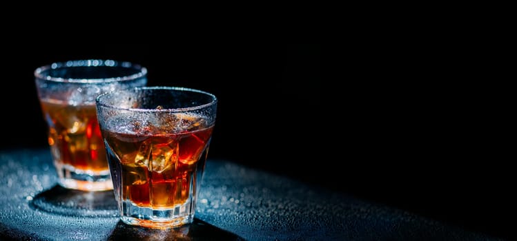 Two glass of cold alcohol cocktail drink on black background with copyspace. Whiskey with ice and cola on bar table. Hard liquor beverage on night club party.