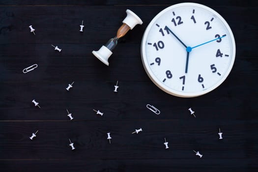 White clock, hourglass and pins on a black wooden background. Top view. Copy space. Education or business concept.