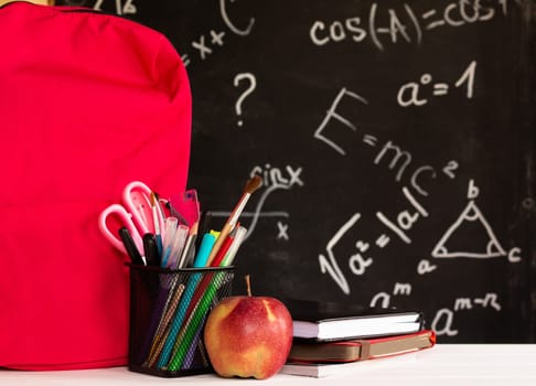 Coloured school supplies, apple, books, backpack on a white contertop on chalkboard background with formulas. Education concept.