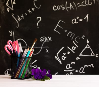 Coloured school supplies and flowers on a white contertop on chalkboard background with formulas. Education concept