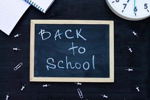 Back to school black chalk board on a black wooden background, white supplies and clock. Top view. Copy space. Education concept.