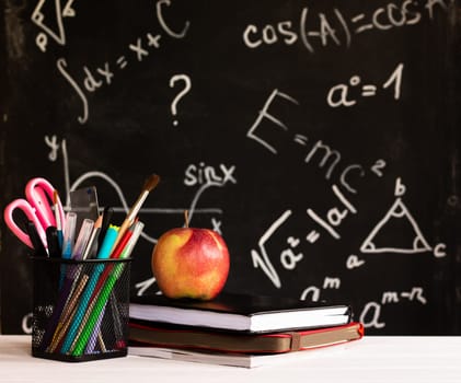 Coloured school supplies, apple, books on a white contertop on chalkboard background with formulas. Education concept