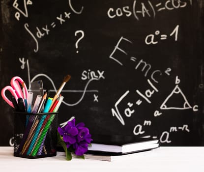Coloured school supplies, flowers, books on white contertop on chalkboard background with formulas. Education concept