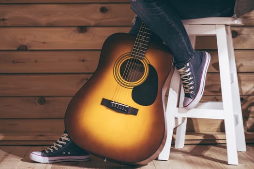 Acoustic six-string yellow-brown guitar and legs of female sitting on stool in jeans, near wooden wall