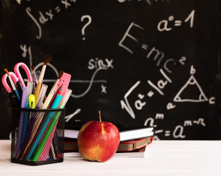 Coloured school supplies, apple, books on a white contertop on chalkboard background with formulas. Education concept