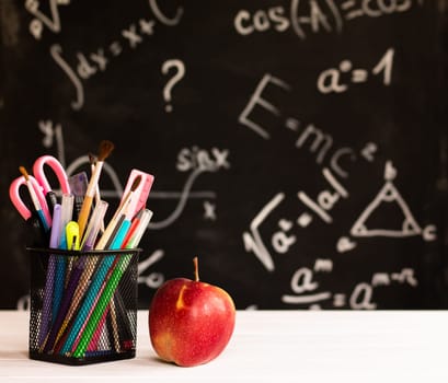 Coloured school supplies and apple on white wooden contertop on chalkboard background with formulas. Education concept.