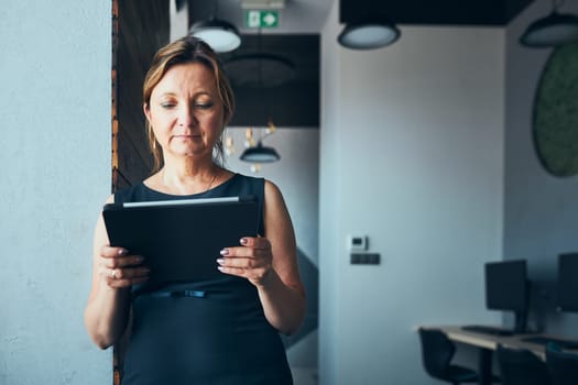 Businesswoman working on tablet in office. Mature woman using touch pad computer standing by window in modern interior. Female manager focused on work holding digital device. Using technology. Open office. Copy space right