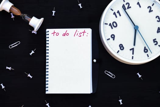 Notebook with To Do List on a black wooden background with clock and hourglass, paper clips. Top view. Copy space