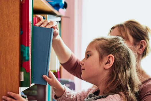 Little girl preschooler choosing a book to read. Child with her mother taking a book from bookshelf
