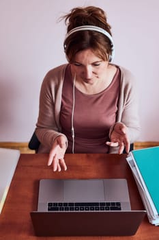 Woman working talking doing her job remotely during video chat phone call on laptop from home. Woman sitting at desk in front of computer looking at screen using headphones and smartphone. Concept of remote working