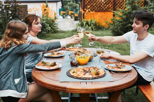 Friends making toast during summer picnic outdoor dinner in a home garden. Close up of people holding wine glasses with white wine over table with pizza, salads and fruits. Dinner in a orchard in a backyard