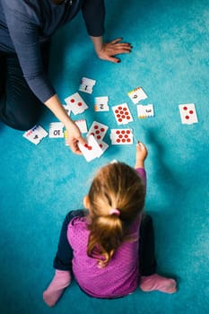 Mother teaching counting her little daughter using card game sitting on a carpet at home