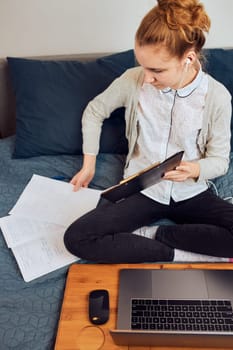 Young woman student having classes, learning online, watching lesson remotely, listening to professor, talking with classmates on video call from home during quarantine. Young girl using laptop, headphones, books, manuals sitting on bed