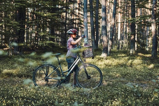 Active woman spending free vacation time on a bicycle trip in a forest on sunny summer day. Woman wearing bicycle helmet and gloves holding bike with basket standing behind the blurred tree leaves