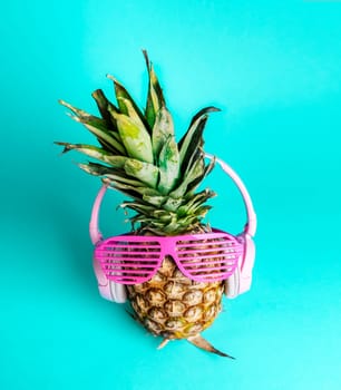 Fashionable trendy pineapple fruit with headphones and sun glasses listen to the music over bright pastel cyan background. Cool hipster ananas with funny face on blue. Summer beach party concept