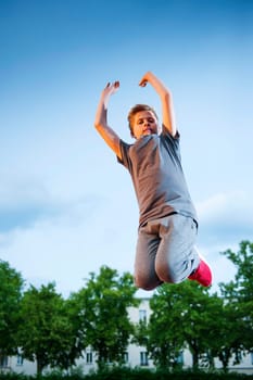 Full body view of a male teenager jumping in the air with his arms stretched up on the sports field against a blue sky.