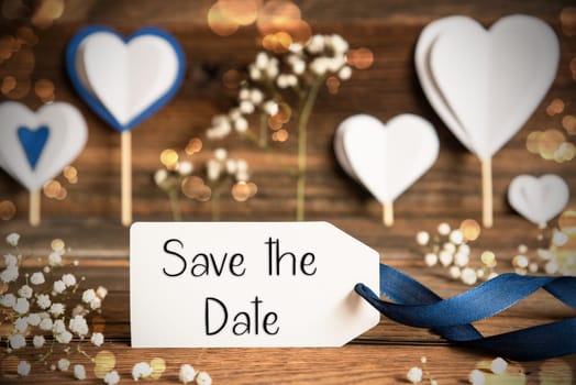 Label With English Text Save The Date. White Festive And Atmospheric Decoration Like Hearts, Flowers And A Blue Bow. Vintage, Wooden Background With Sparkling Light.