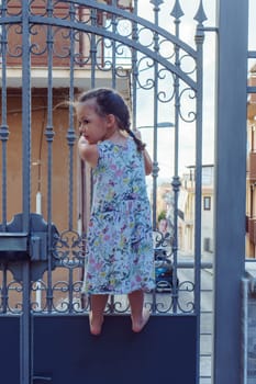 Vertical curious, cute, barefoot blonde little girl climbing on metal fence, gate in urban city area against houses and streets. Home arrest, stay at home and be safe. Children protection and care