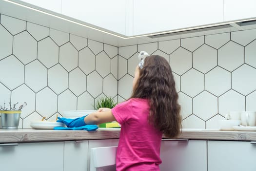 Back view of little preteen girl with loose long dark hair wearing pink T-shirt, taking off blue rubber gloves, standing near table after washing dishes near plates in kitchen. Housework, dishwashing.