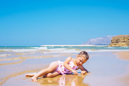 Cute little girl in pink swimsuit with strawberries lying in the sand and playing with water at the seashore against blue sky. Joying low tide and warm summer. Learn to swim on vacation. Copy space