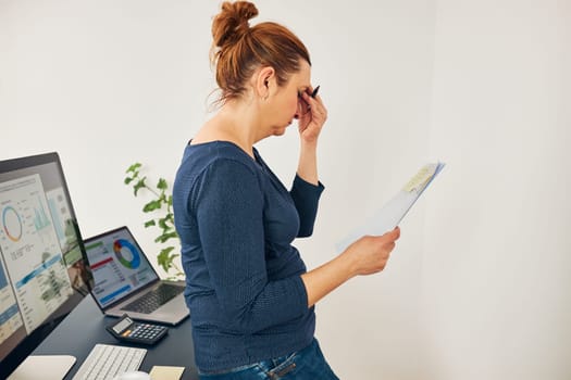 Woman entrepreneur feeling tired and stressed. Puzzled and confused businesswoman thinking hard holding doccuments standing at her desk in office. Frustrated woman keeping eyes closed focused on solving difficult work