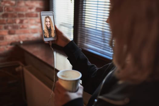 Young woman having video call, talking remotely, taking selfie photo holding smartphone. Girl drinking coffee relaxing while taking break in office standing at window