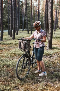 Active woman spending free summer vacation time on a bicycle trip in a forest. Woman wearing bicycle helmet and gloves holding bike with basket