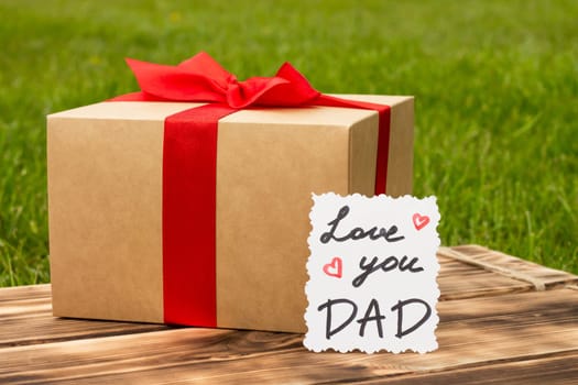 Greeting card Love You Dad with a gift box on a rustic wooden tabletop and green grass background