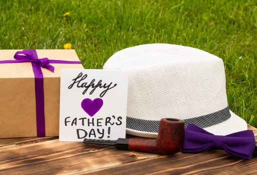 Greeting card Happy Father's Day with a gift box and men's accessories on a rustic wooden tabletop and grass background