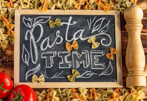 Chalk board sign Pasta Time on a wooden tabletop with raw farfalle, fresh tomatoes and black pepper.