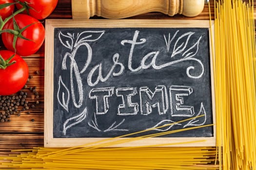 Chalk board sign Pasta Time on a wooden tabletop with raw spaghetti, fresh tomatoes and black pepper.