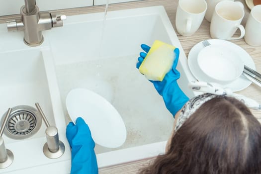 Unrecognizable little preteen girl wearing big blue rubber gloves, standing near sink, holding yellow soapy sponge, washing white plate in kitchen. Housekeeping, housework, dishwashing. Top view.