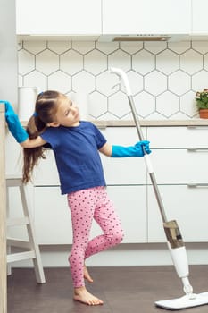 Portrait of happy little preteen girl with long dark hair wearing blue T-shirt, pink leggings, big blue rubber gloves, holding handheld spray mop, leaning on table in kitchen. Housework, housekeeping.