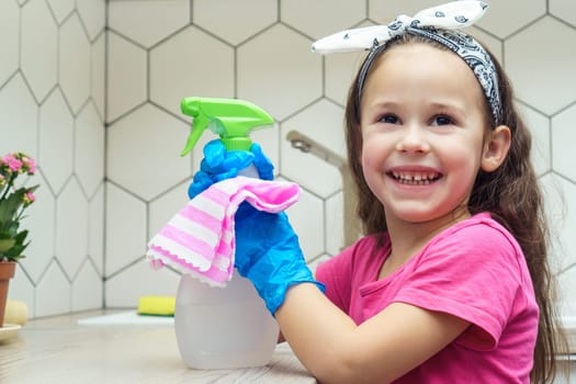 Portrait of cute little preteen girl with dark hair wearing pink T-shirt, big blue rubber gloves, holding cleanser sprayer, rag on white table for cleaning washing in kitchen. Housework, housekeeping.