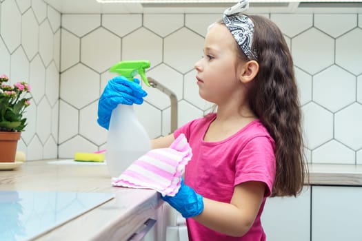 Side view of concentrated little preteen girl with long dark hair wearing pink T-shirt, big blue rubber gloves, looking at spray bottle, holding rag near table in kitchen. Housework, housekeeping.