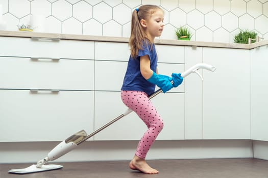 Portrait of funny little preteen girl with long dark hair wearing blue T-shirt, pink leggings, blue rubber gloves, riding on handheld spray mop as witch near table in kitchen. Housework, housekeeping.