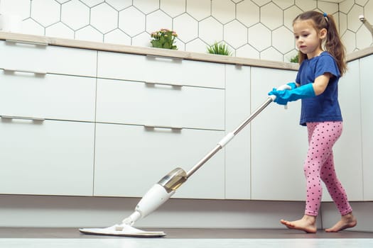 Side view of cute little preteen girl with long dark hair wearing blue T-shirt, pink leggings, big blue rubber gloves, holding microfiber spray mop, cleaning floor in kitchen. Housework, housekeeping.