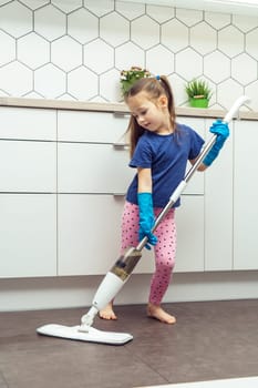 Portrait of busy little preteen girl with long dark hair wearing blue T-shirt, pink leggings, big blue rubber gloves, holding handheld spray mop, washing floor in kitchen. Housework, housekeeping.