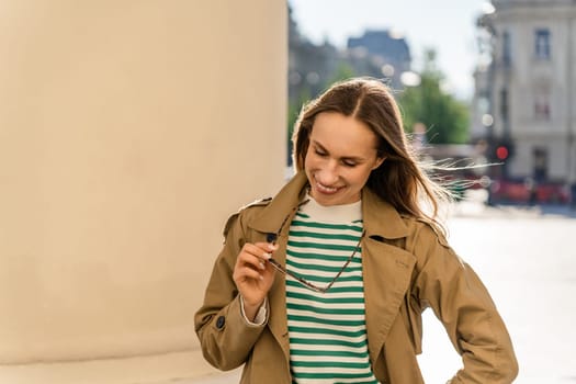 A woman smiles and holds sunglasses while walking in the city.