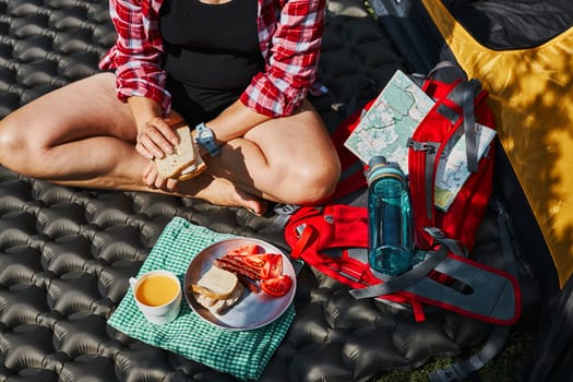 Woman having breakfast sitting by tent at camping. Female actively spending summer vacations close to nature outdoors. Concept of camp life