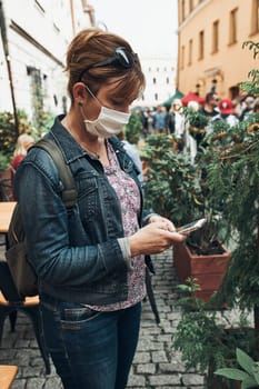 Young woman using smartphone social media sending texts watching photos and videos talking remotely with friends standing in a crowded street downtown. Woman is wearing the face mask to avoid virus infection and to prevent the spread of disease in time of coronavirus