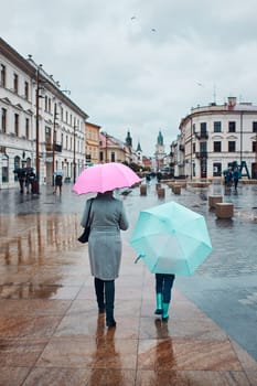 Back view of mother and her daughter holding the pink and blue umbrellas walking in a downtown on rainy gloomy autumn day