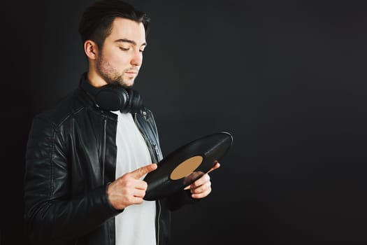 Handsome man looking at vinyl record. Rock style. Vintage music style. Male wearing black jacket holding turntable plate standing on black background. Retro music. Classic audio. Copy space right
