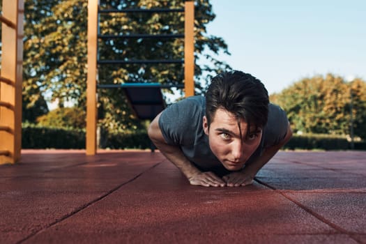 Young man doing push-ups on a red rubber ground during his workout in a modern calisthenics street workout park. Man wearing sportswear looking away