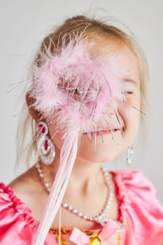 Little girl enjoying her role of princess. Adorable cute 5-6 years old girl wearing pink princess dress holding magic wand at front of her face standing on plain background