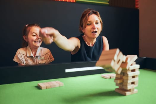 Excited woman playing jenga game with her daughter in play room. Woman removing block from stack. Falling tower of wooden blocks. Game of skill and fun. Family time