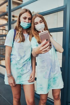 Young women girls taking selfie photos using smartphone standing at store front downtown. Girls wearing face masks to avoid virus infection and to prevent the spread of disease in time of coronavirus