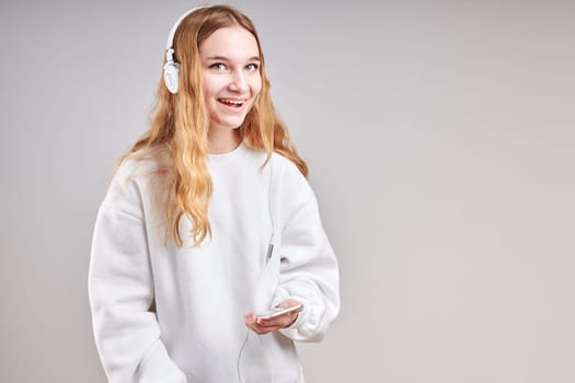 Young woman girl listening to music streaming content having fun watching video enjoying video chat talking with friends making gestures faces using smartphone earphones headphones standing over plain grey background