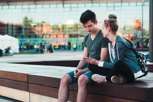 Couple of friends, teenage girl and boy,  having fun together with smartphones,  sitting in center of town, spending time together