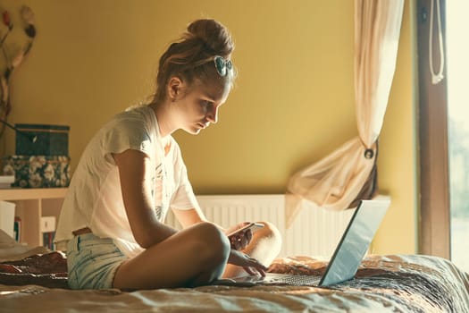 Young woman using portable computer and mobile phone, sitting on a bed, learning online at home. Candid people, real moments, authentic situations